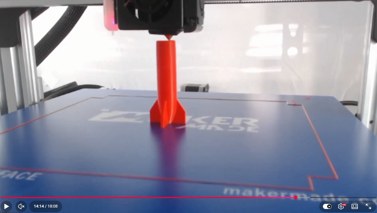 A bright red 3D-printed rocket model being printed on a blue Maker Faire branded 3D printer bed by an overhead printer arm.